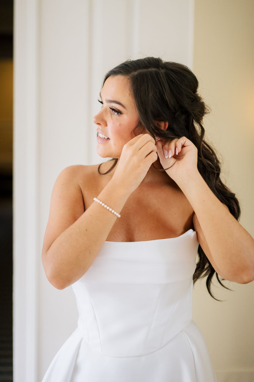 Someone in white strapless dress putting on jewelry while getting ready for special occasion.