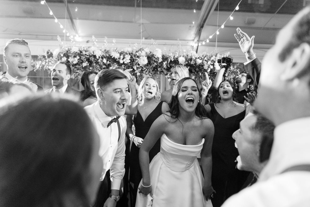 An energetic crowd of wedding reception guests dancing and celebrating with raised arms.