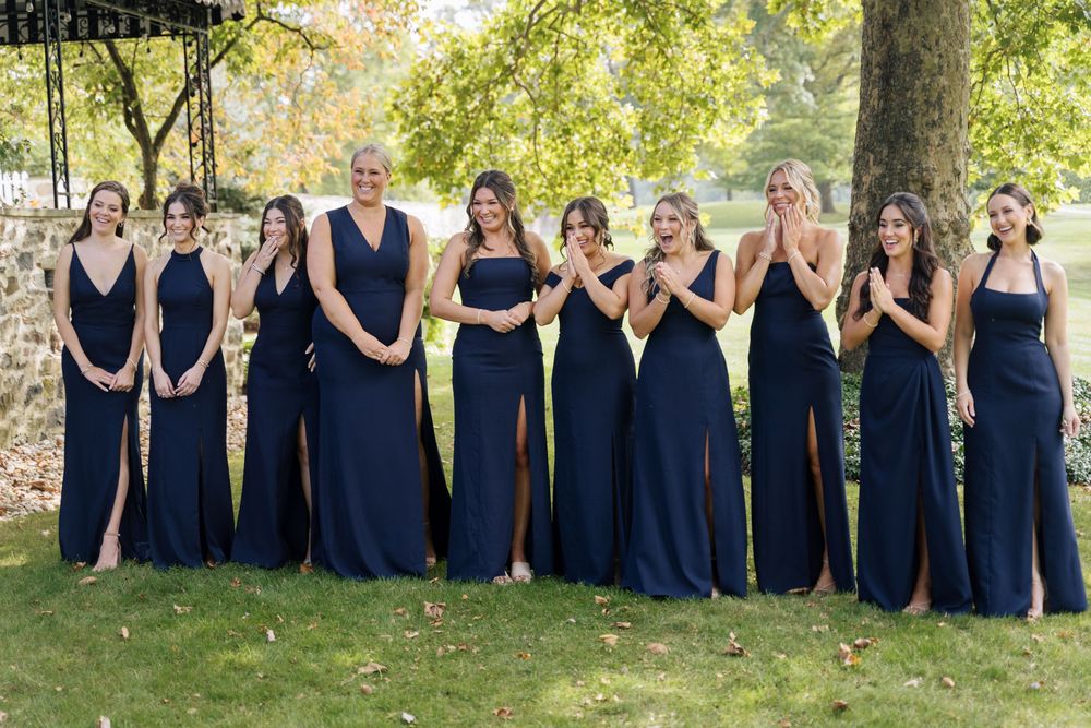 Wedding party poses outdoors wearing matching navy blue dresses against a sunny backdrop.