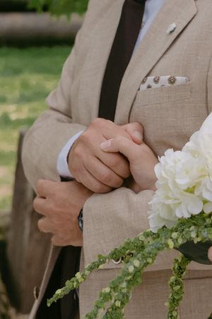 Close-up details of hands holding white floral bouquet against beige suit.