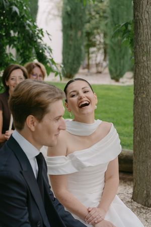A joyful wedding moment captured outdoors with a couple sitting together under green trees.
