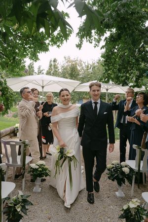 Wedding party walking together on gravel path under trees with white umbrellas.