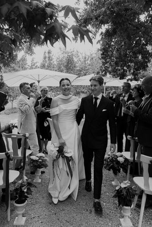 Black and white photo of wedding celebration with guests walking outdoors under trees.