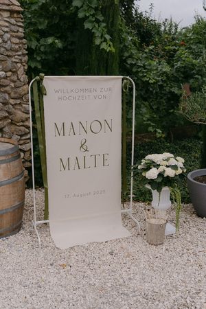 Wedding welcome sign with names displayed on white gravel path with stone wall backdrop.