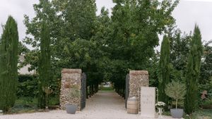 Tree-lined pathway with stone pillars and wine barrels in an outdoor garden setting.