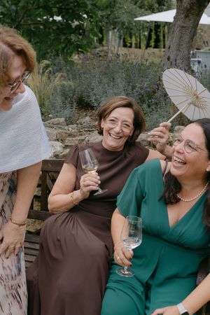 Two wedding guests in brown and green dresses share a joyful moment while holding decorative fans at an outdoor celebration.