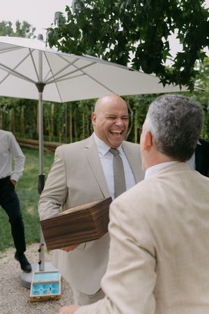 Two guests in light colored suits share a laugh while holding wooden boxes at outdoor reception.
