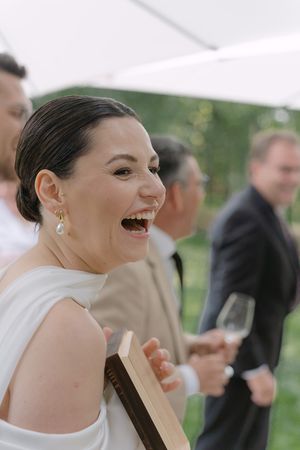 A bride in a white dress laughs joyfully during an outdoor wedding celebration.