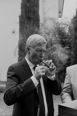 A gentleman in a suit lights a cigar outdoors with cypress trees in the background.