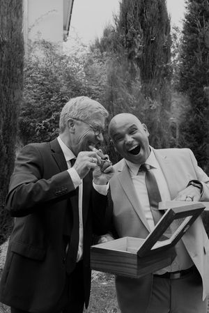 Two wedding guests share a lighthearted moment while lighting cigars in a garden setting.