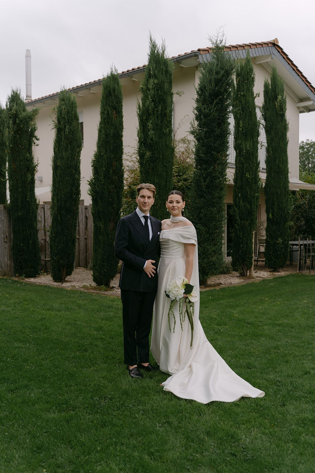 Elegant wedding couple poses in front of Mediterranean-style house with tall cypress trees.