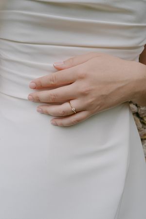 Close-up of delicate wedding ring on bride's hand against white dress fabric.