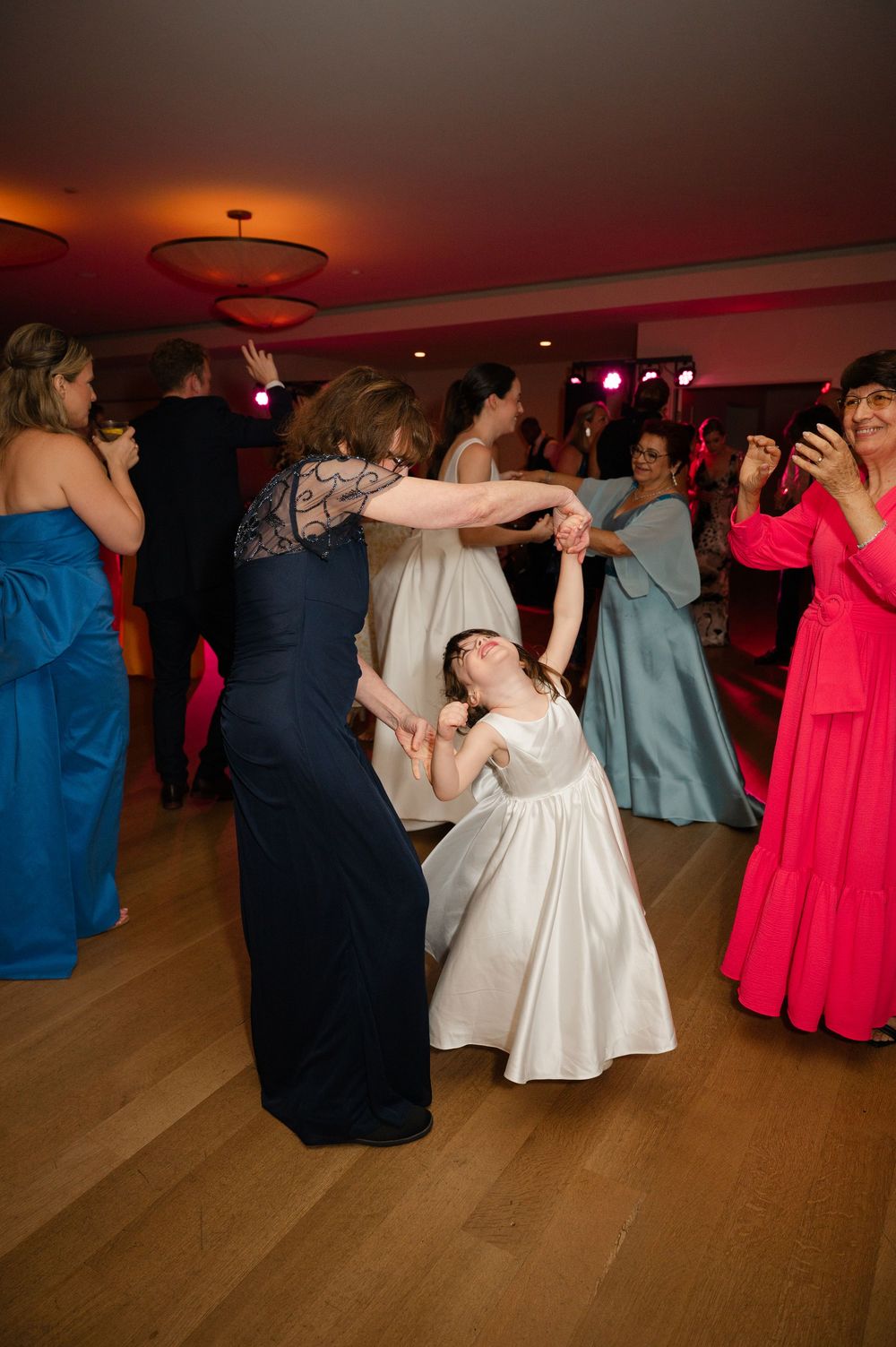 Wedding guests dance and twirl on a wooden floor at reception with colorful formal dresses and ambient lighting.