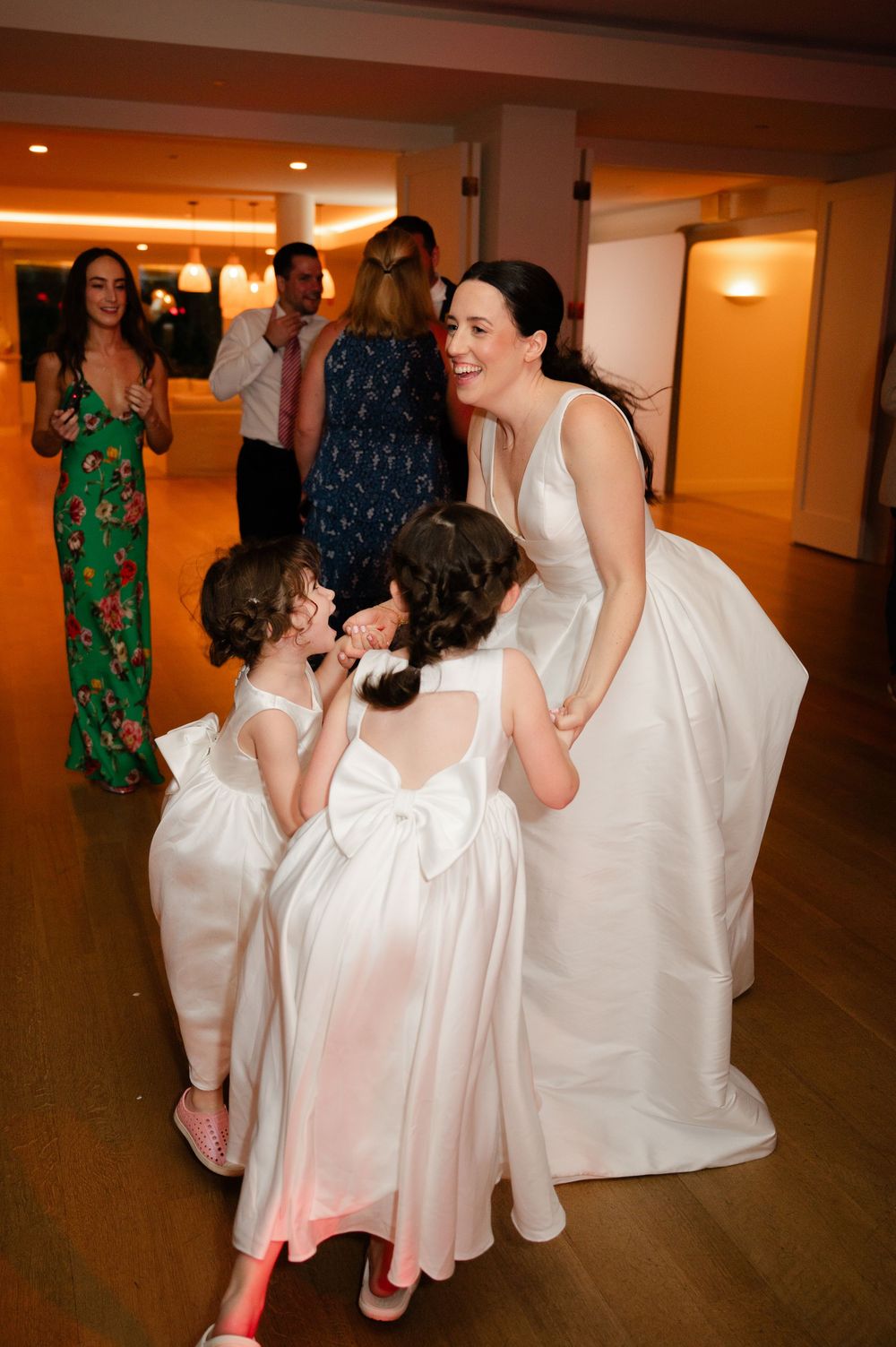 A group of people dance and celebrate in a warm-lit ballroom wearing white and floral attire.