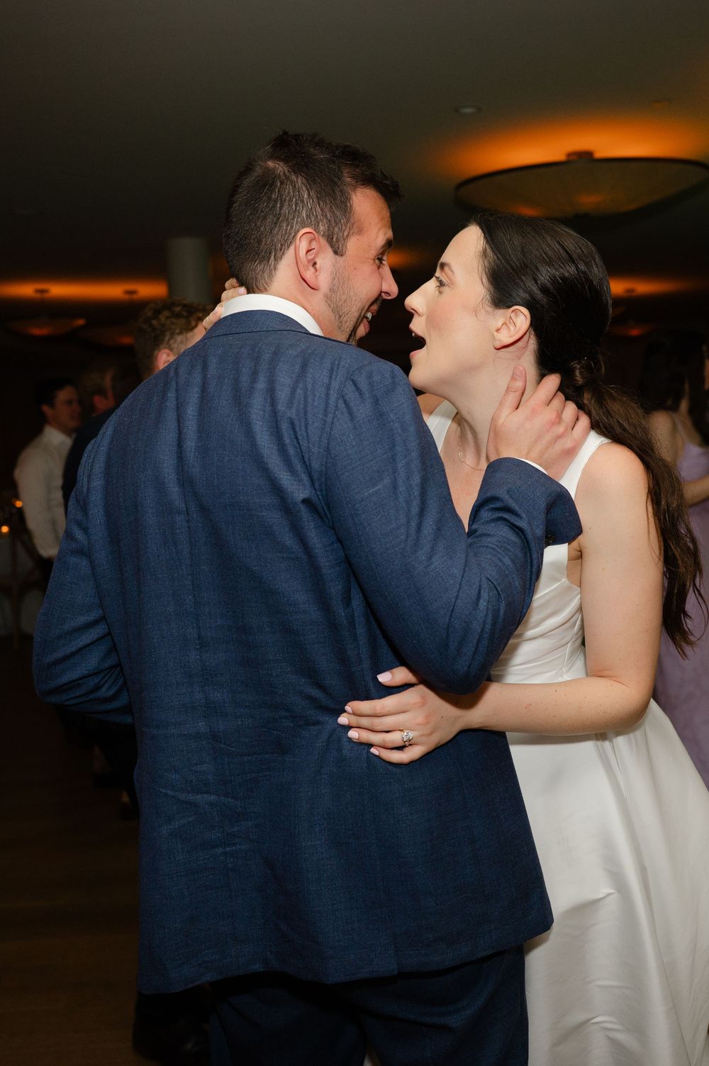 A couple in formal wedding attire share their first dance together in front of guests at a dimly lit reception.