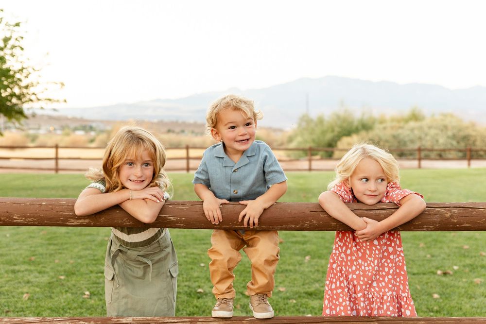 3 kids hanging on a wood fence looking at mom and dad. 2 year old boy in the middle with a sister on either side