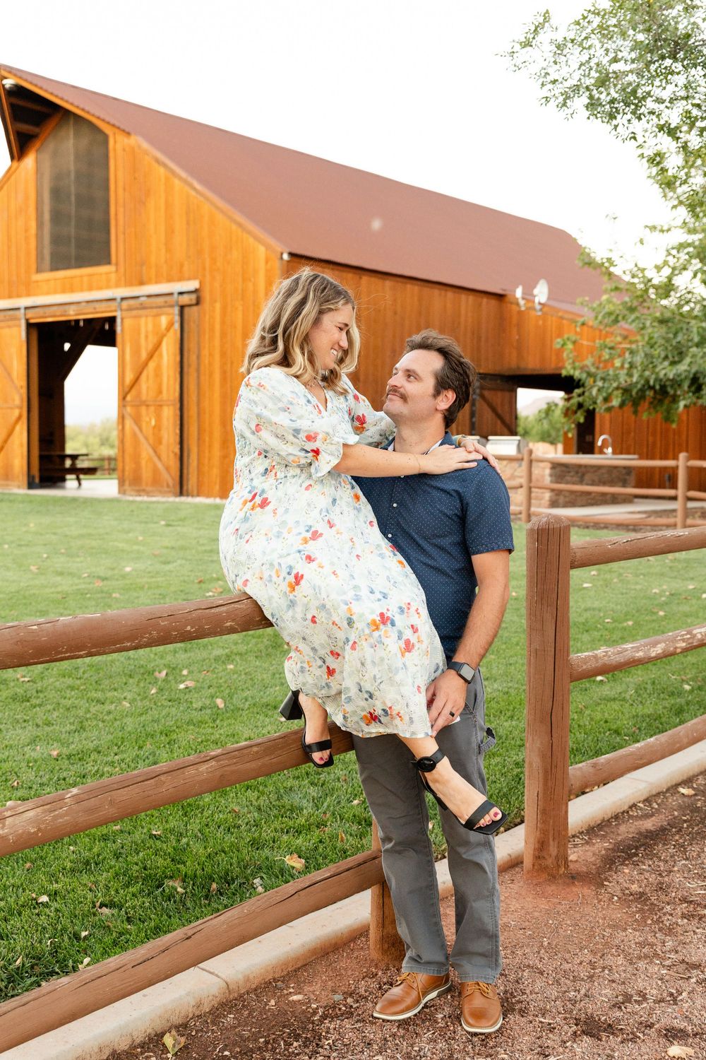 st. George utah woman in a floral dress sitting on top of a wood farm fence. arms around husband kicking one foot out smiling at each other