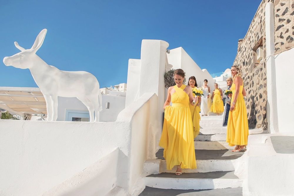 Women in yellow dresses stand on white stairs beside a large white sculpture of a deer in Santorini, Greece.
