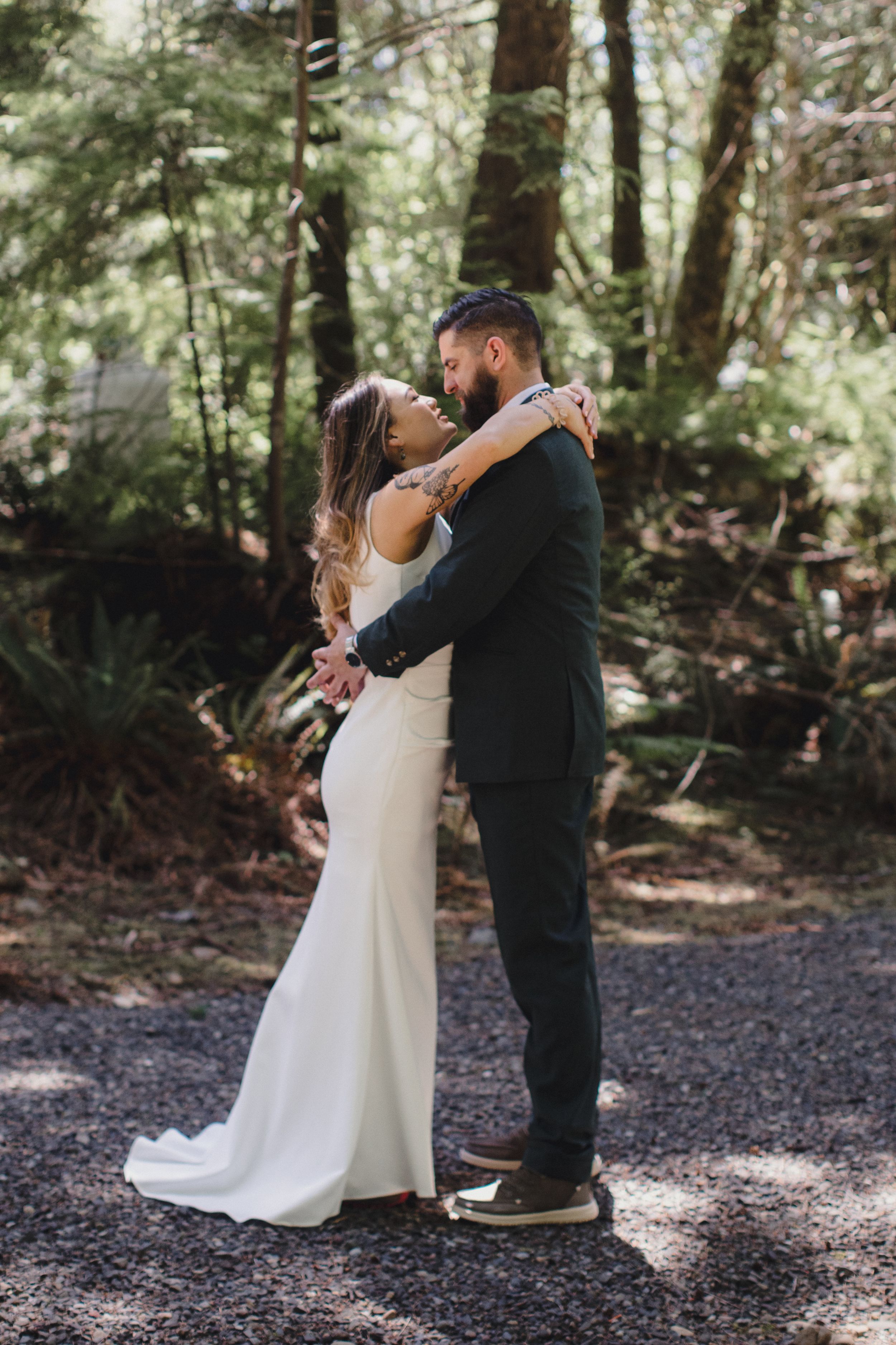 A couple embraces in a romantic forest wedding photo, with the bride in a sleek white dress and groom in dark suit.