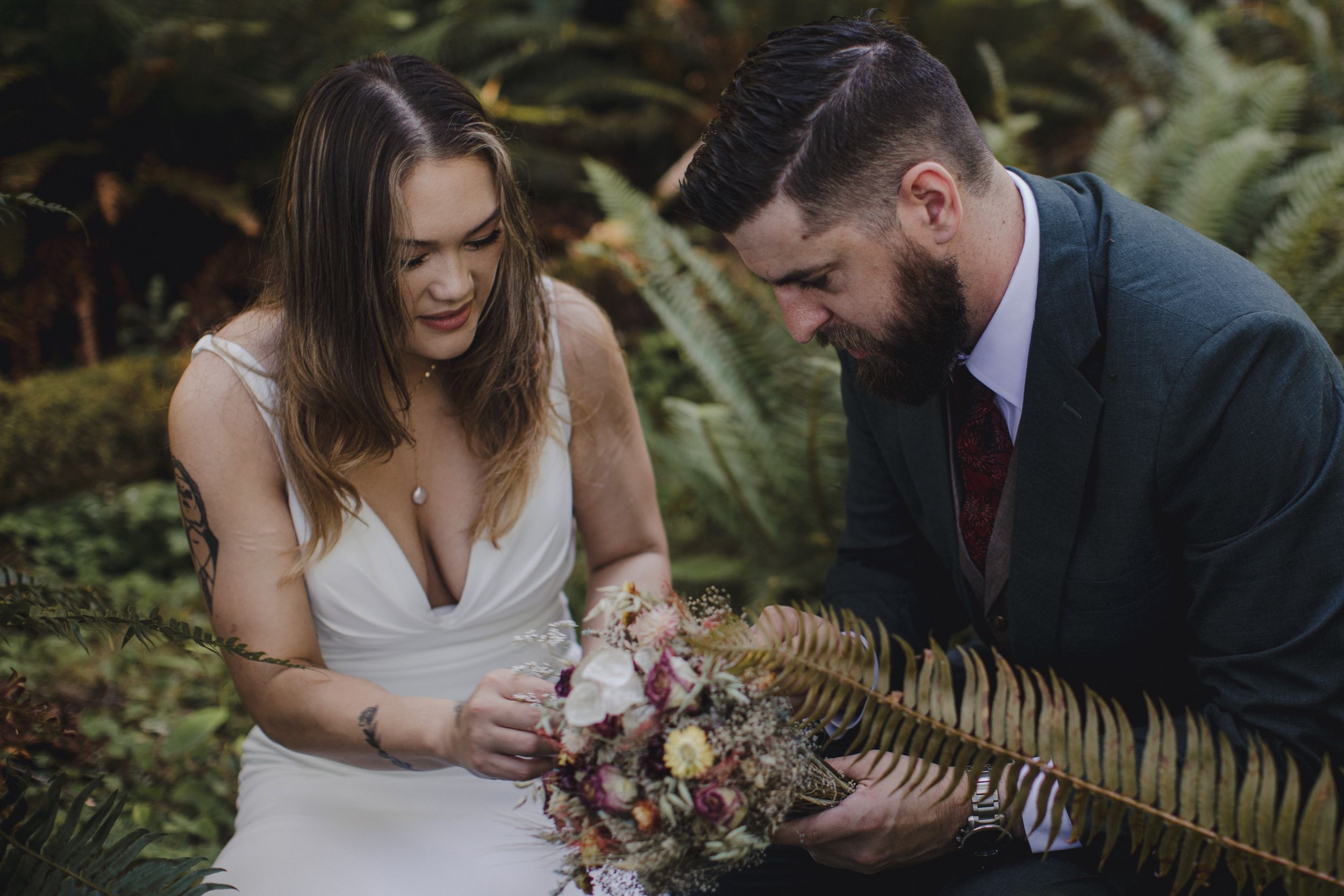 A couple shares an intimate moment in a lush garden setting while looking at a rustic bouquet of flowers together.