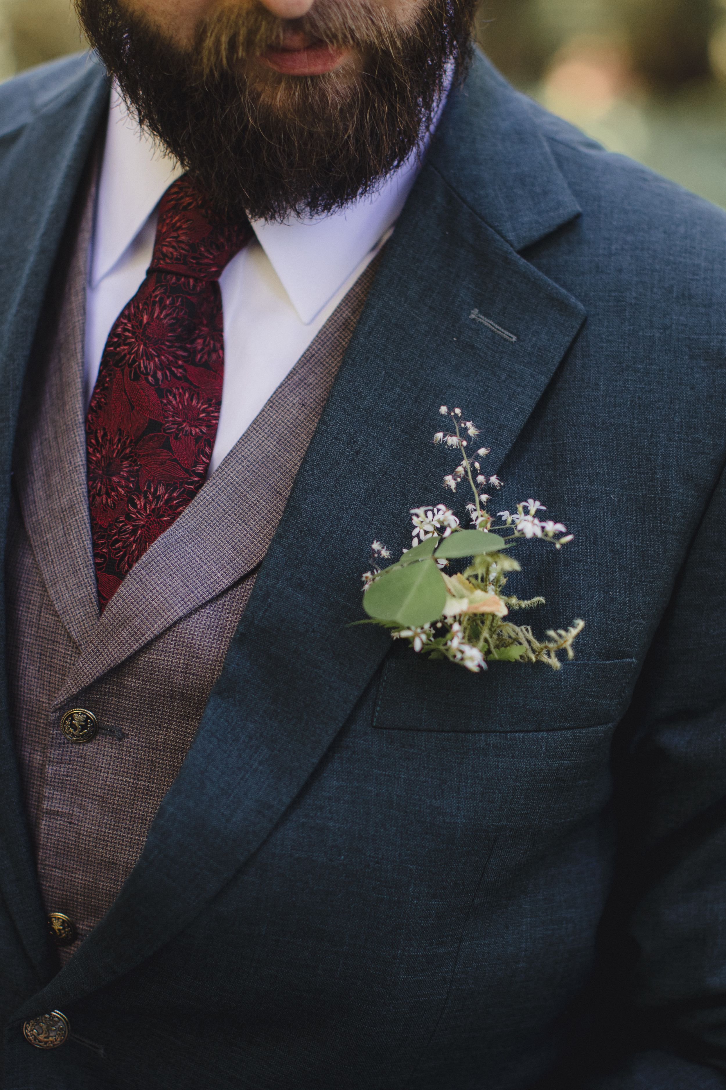 A close-up of a navy suit jacket with brown vest and red tie, adorned with a delicate floral boutonniere.