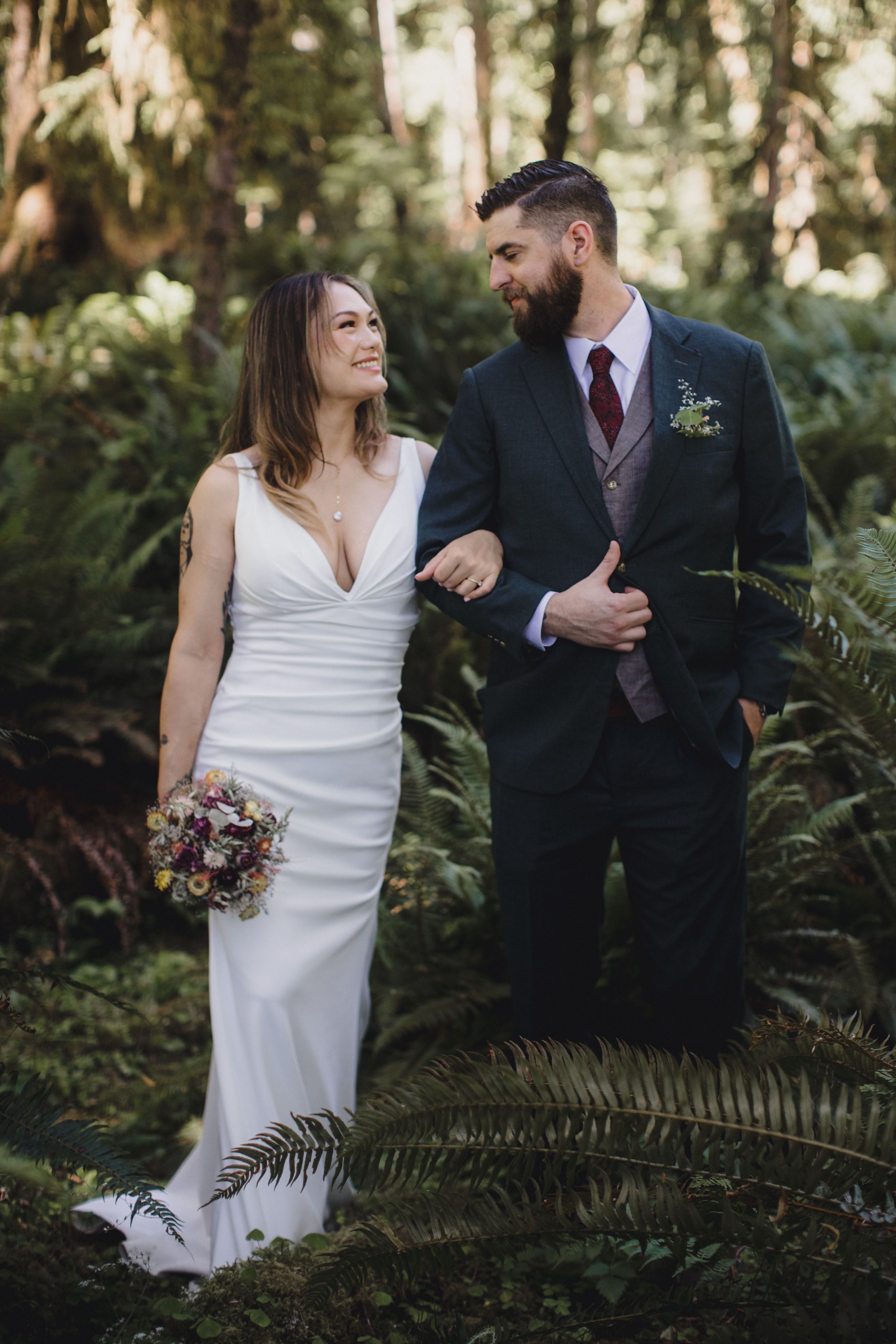 A bride in a fitted white dress and groom in dark suit share a romantic moment among ferns in a forest setting.
