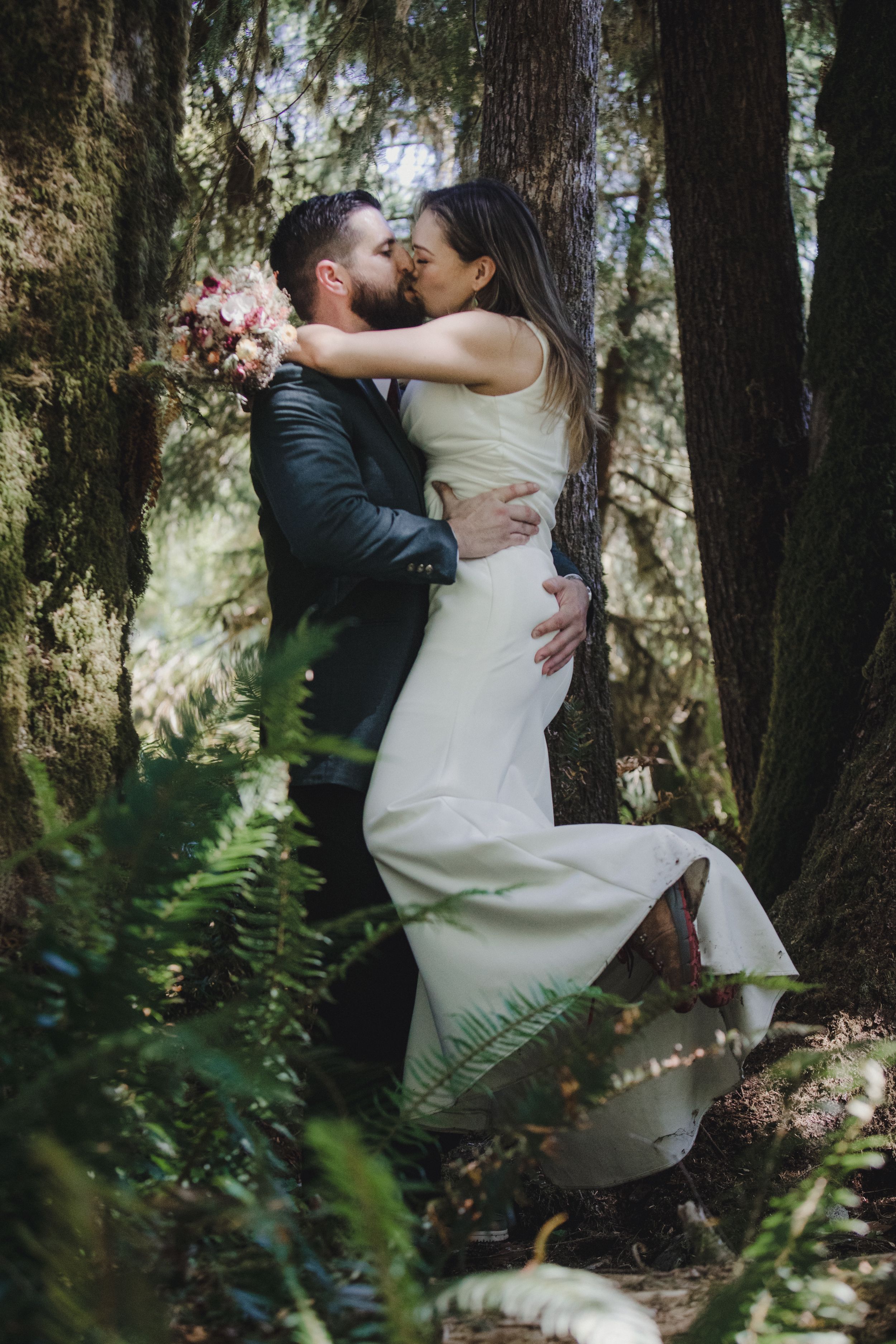 A couple shares an intimate embrace against a tree trunk in a lush forest setting during their rustic outdoor wedding photoshoot.