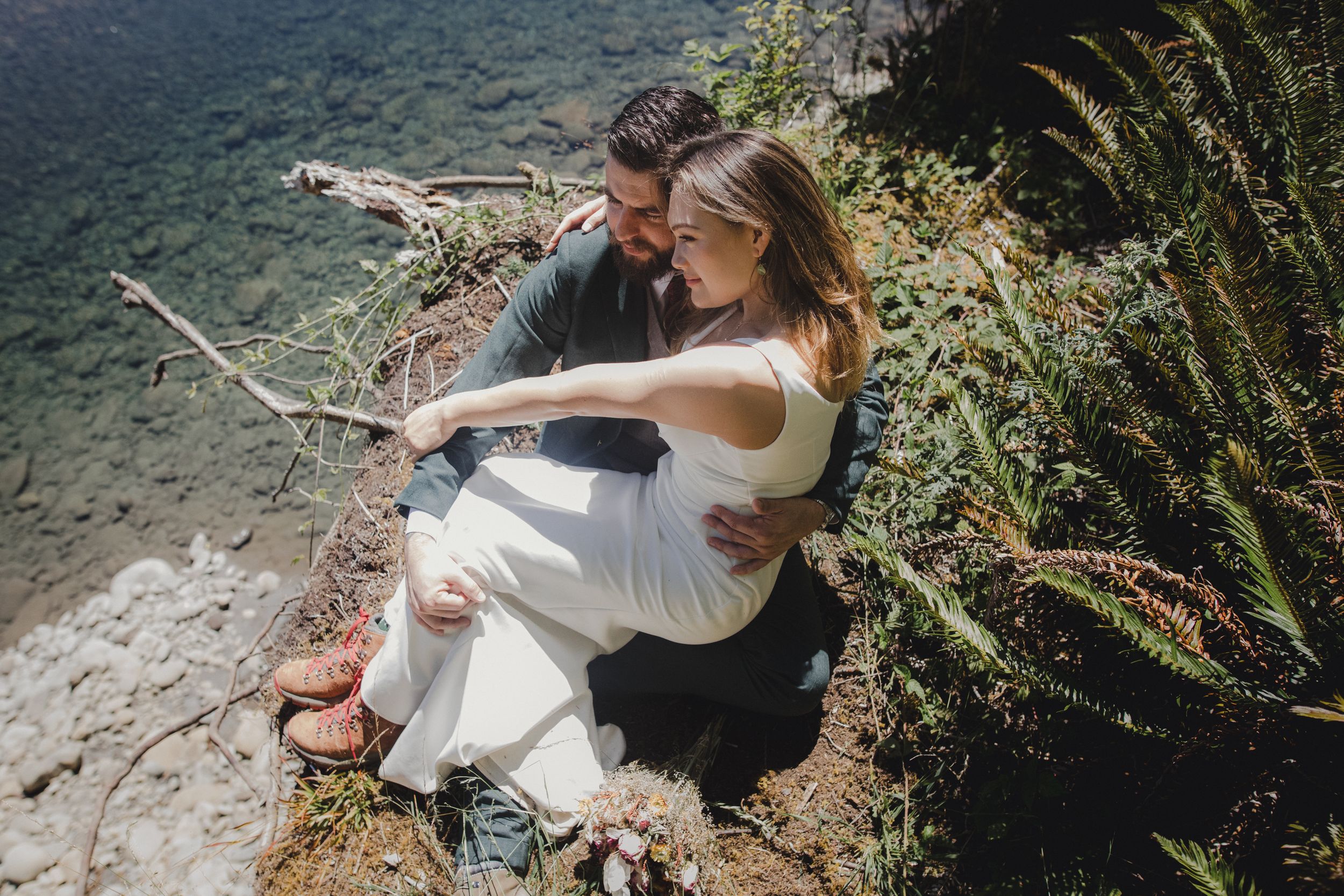 A couple shares a tender moment while sitting on a cliff edge overlooking turquoise waters and coastal scenery.