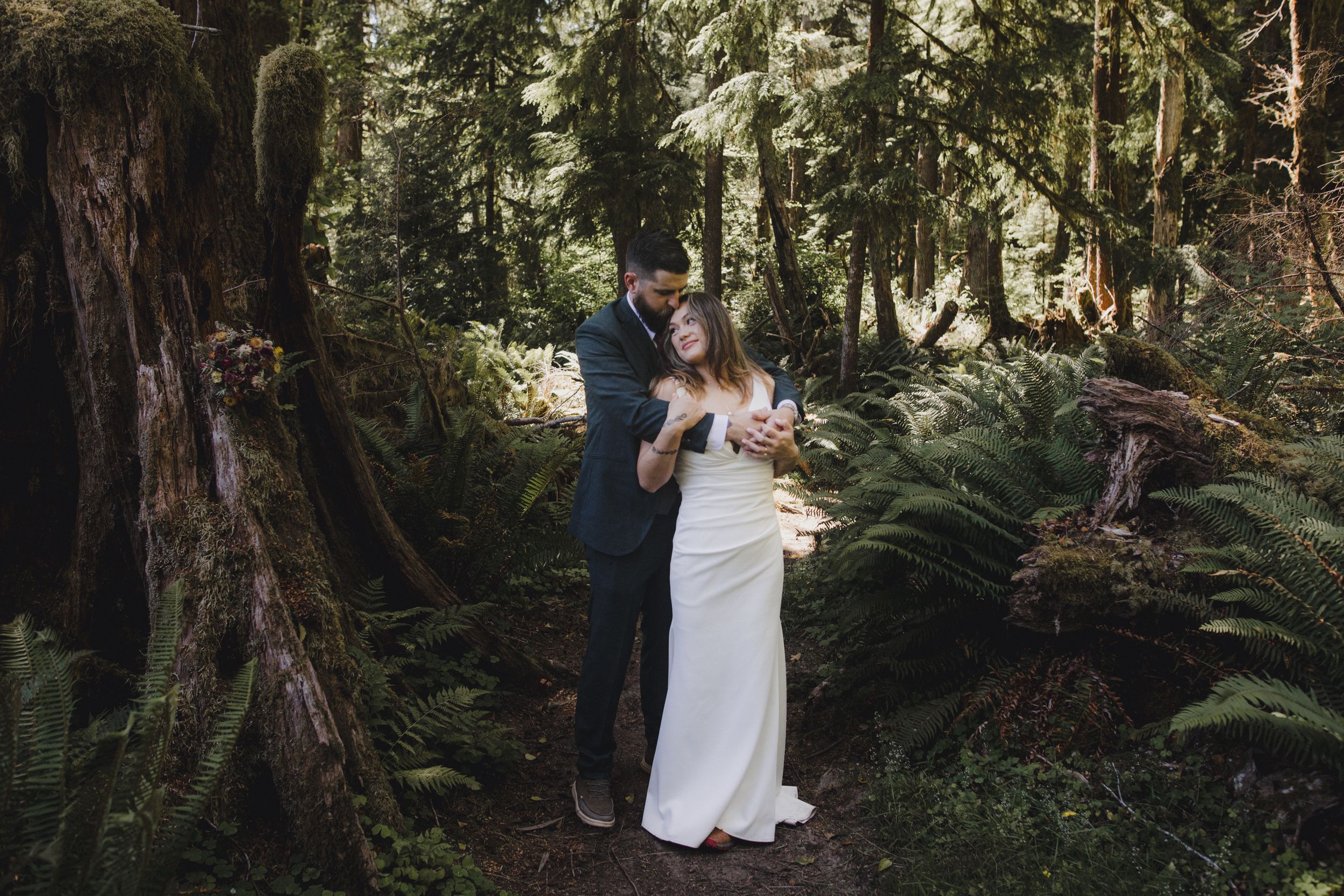 A couple in wedding attire embraces in a lush forest setting surrounded by towering trees and ferns.
