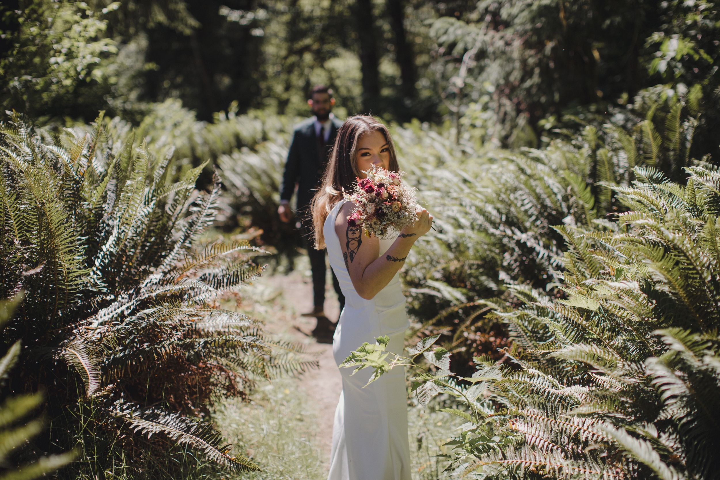 A bride in a sleek white dress poses in a sunlit garden path surrounded by lush ferns.