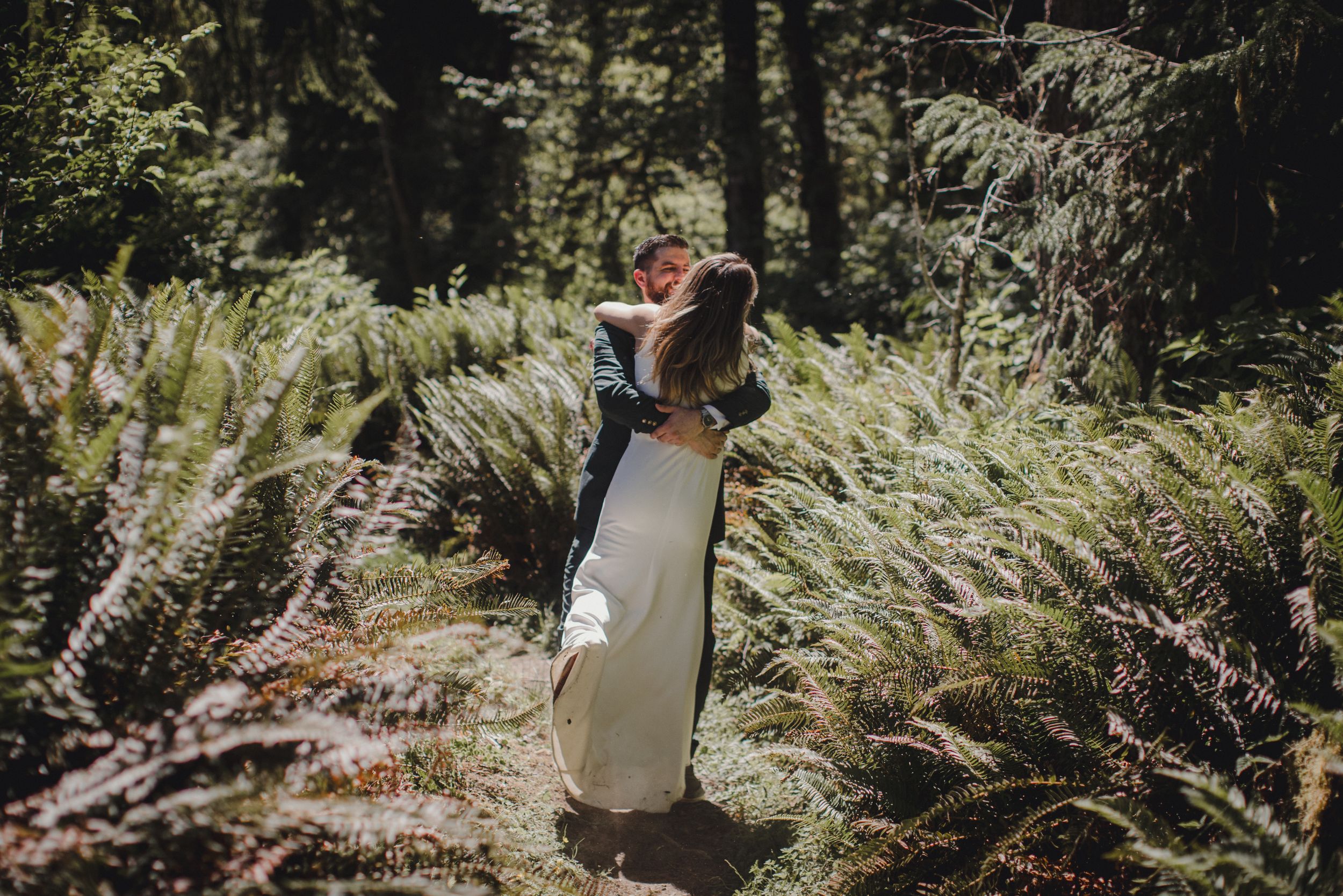 A wedding couple shares an intimate moment surrounded by ferns in a forest setting.