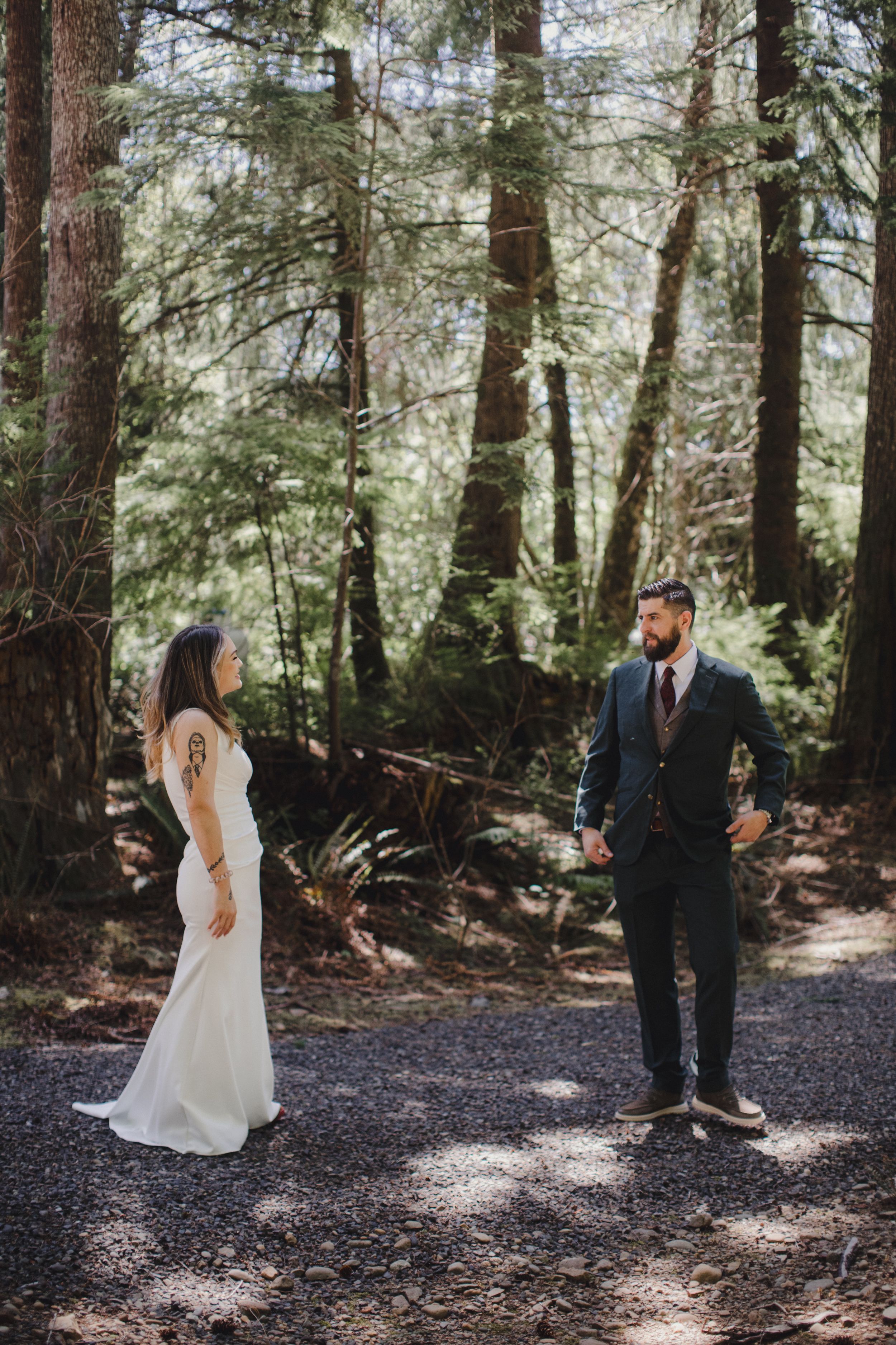 A couple in wedding attire stands facing each other on a forest path surrounded by tall evergreen trees and dappled sunlight.