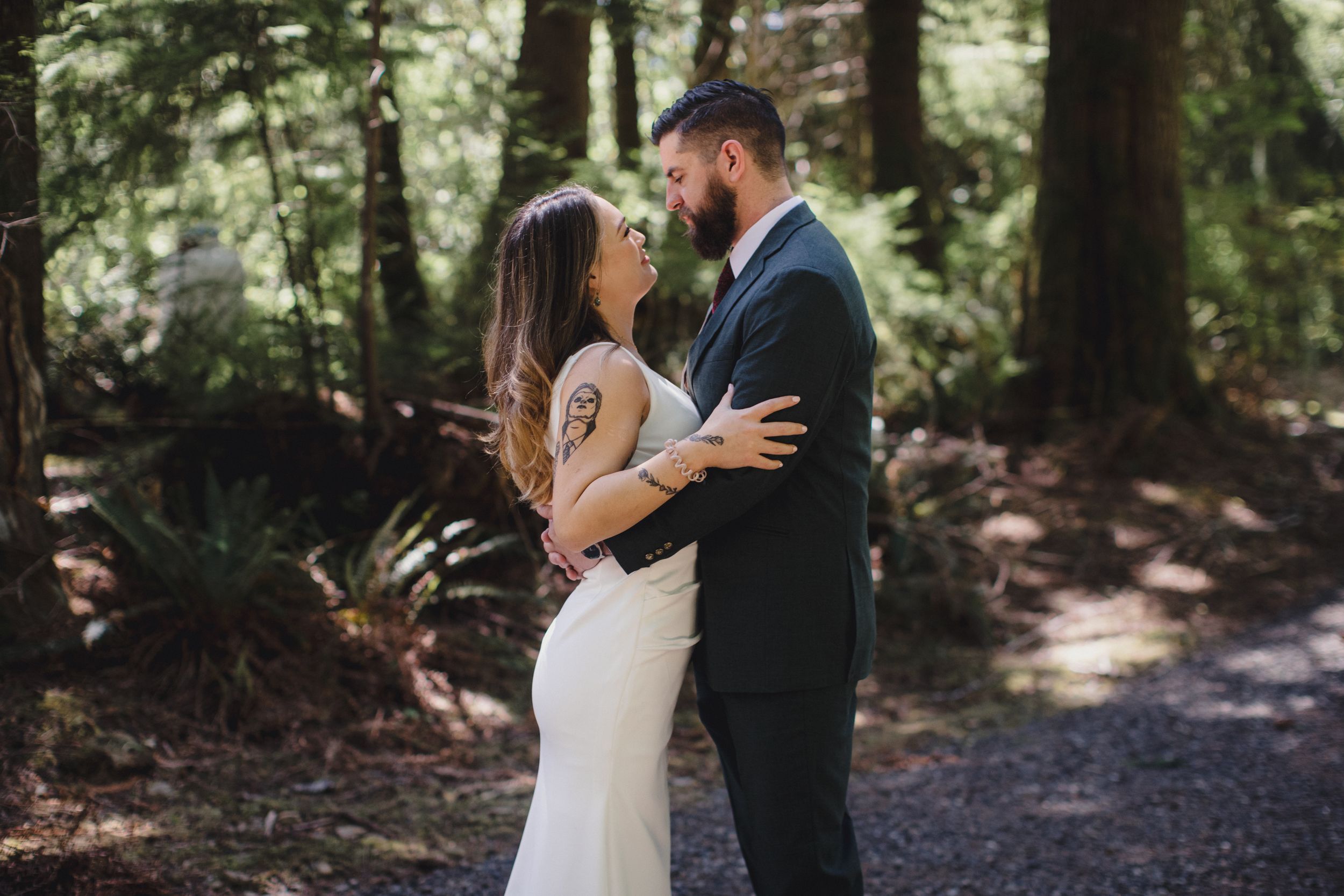 A loving couple shares an intimate embrace on a sunlit forest path surrounded by tall trees and dappled light.