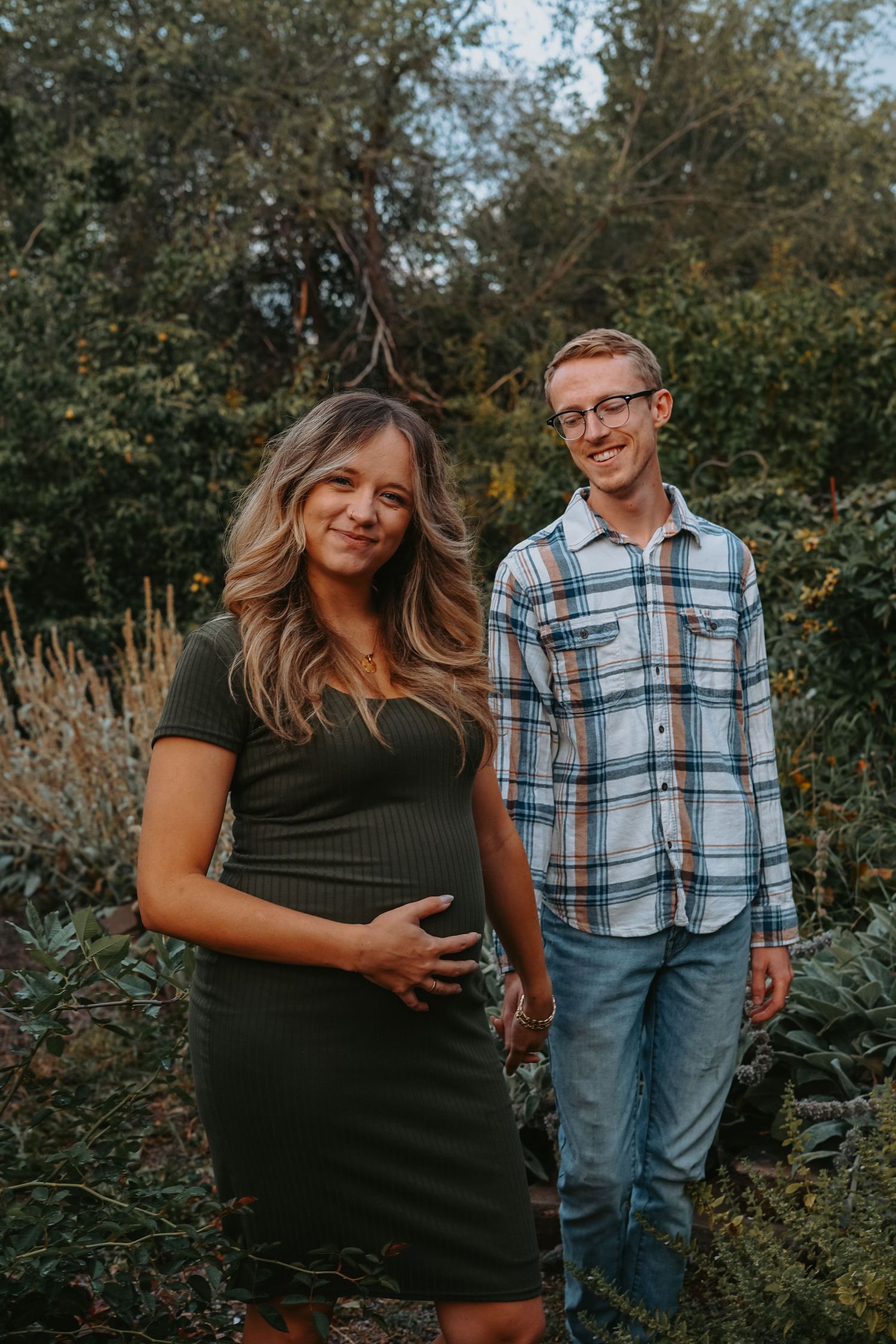 Couple poses together in outdoor garden setting with lush greenery and trees in the background during golden hour.
