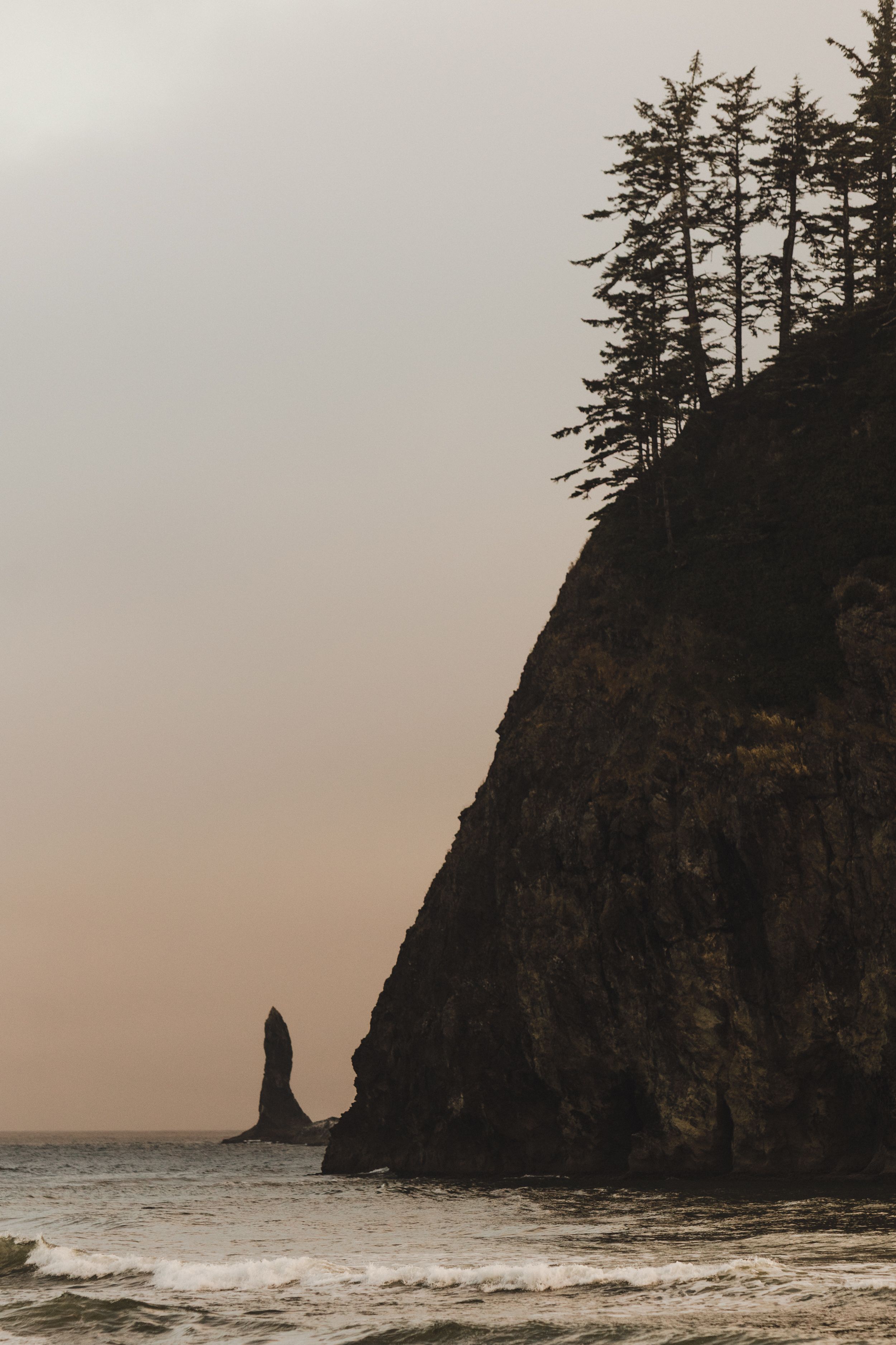 Majestic sea stack and rugged cliffside with evergreen trees silhouetted against moody sky at coastal beach.