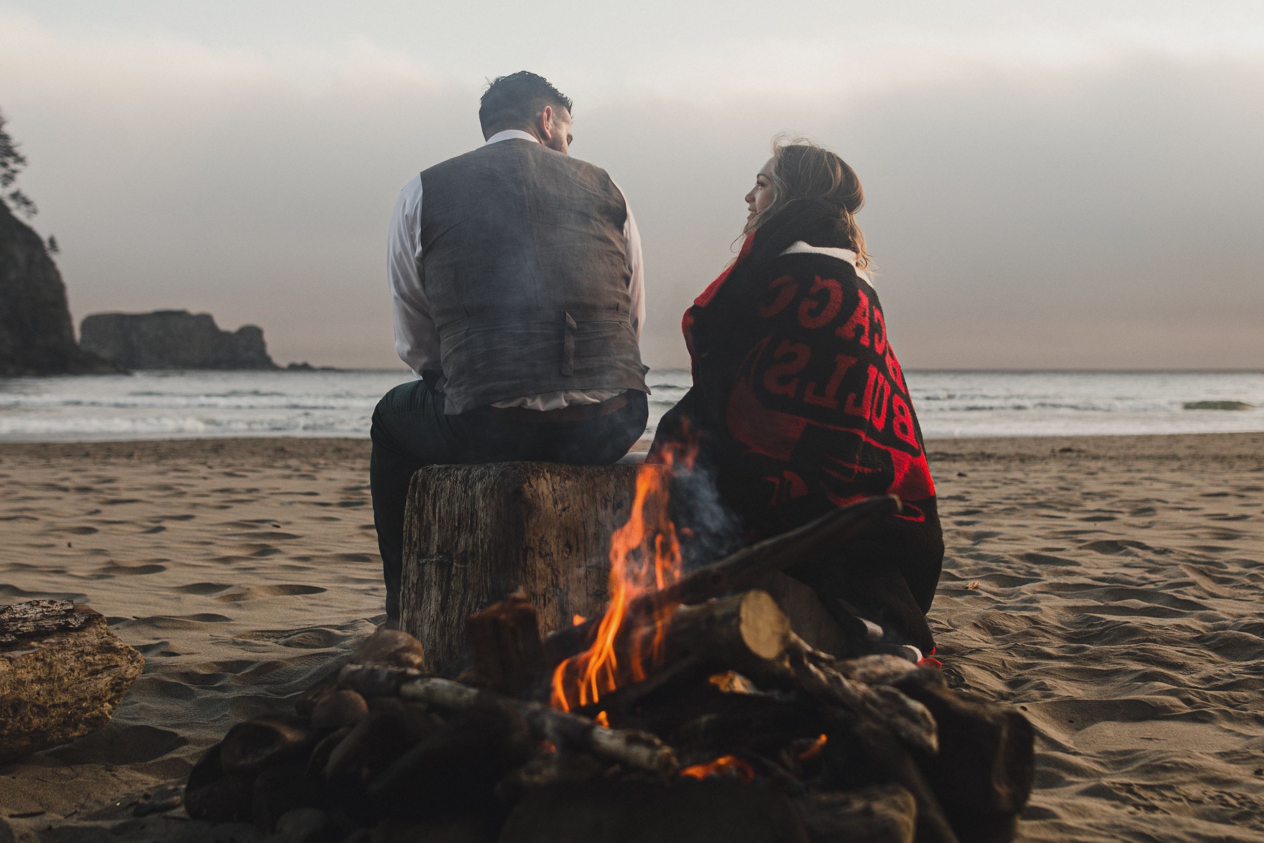 An eloping couple sits together on the beach near a warm bonfire as waves break on the shore at sunset.