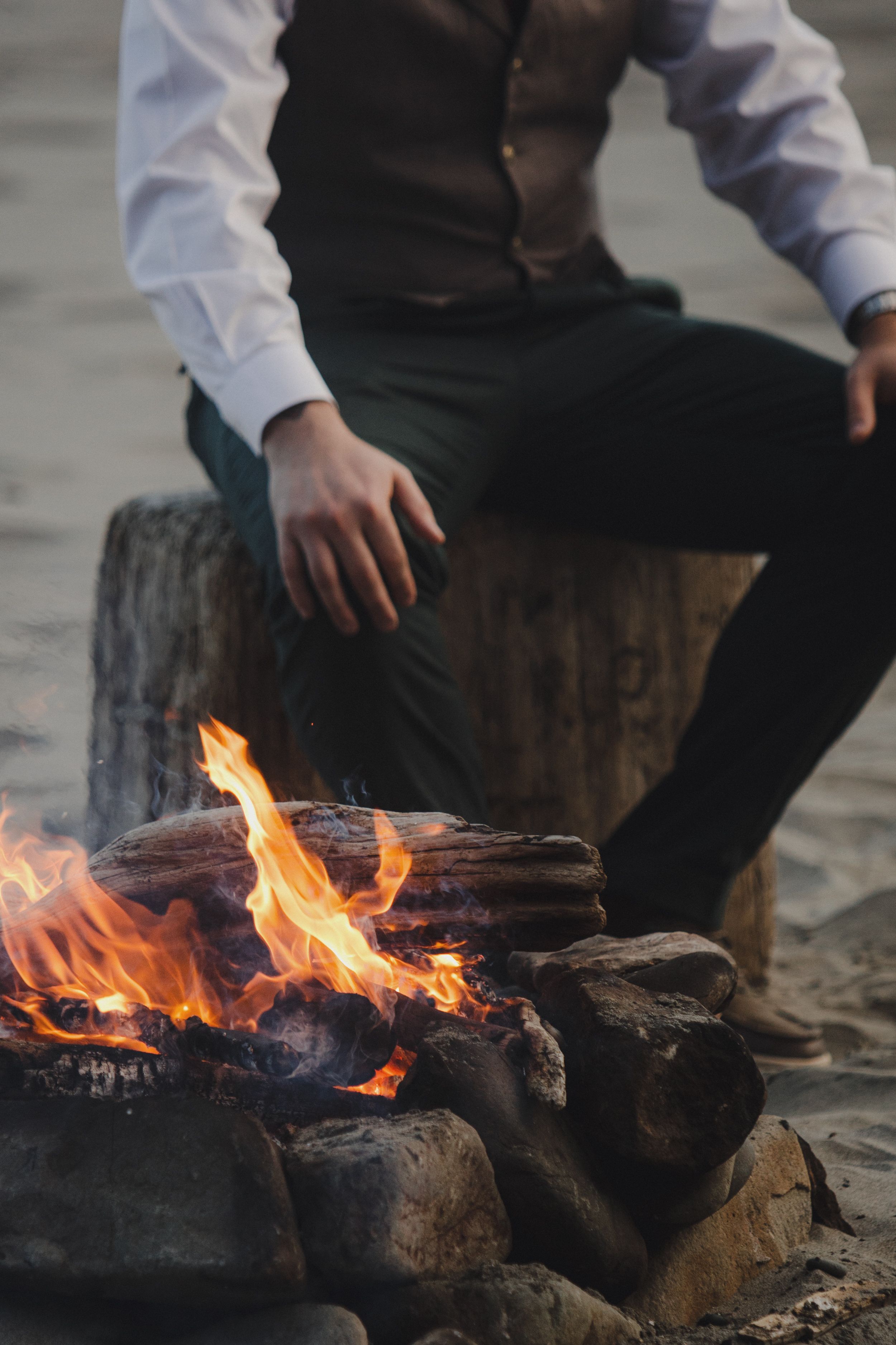 A cozy campfire burns among rocks on a beach while someone sits nearby on a log during sunset.