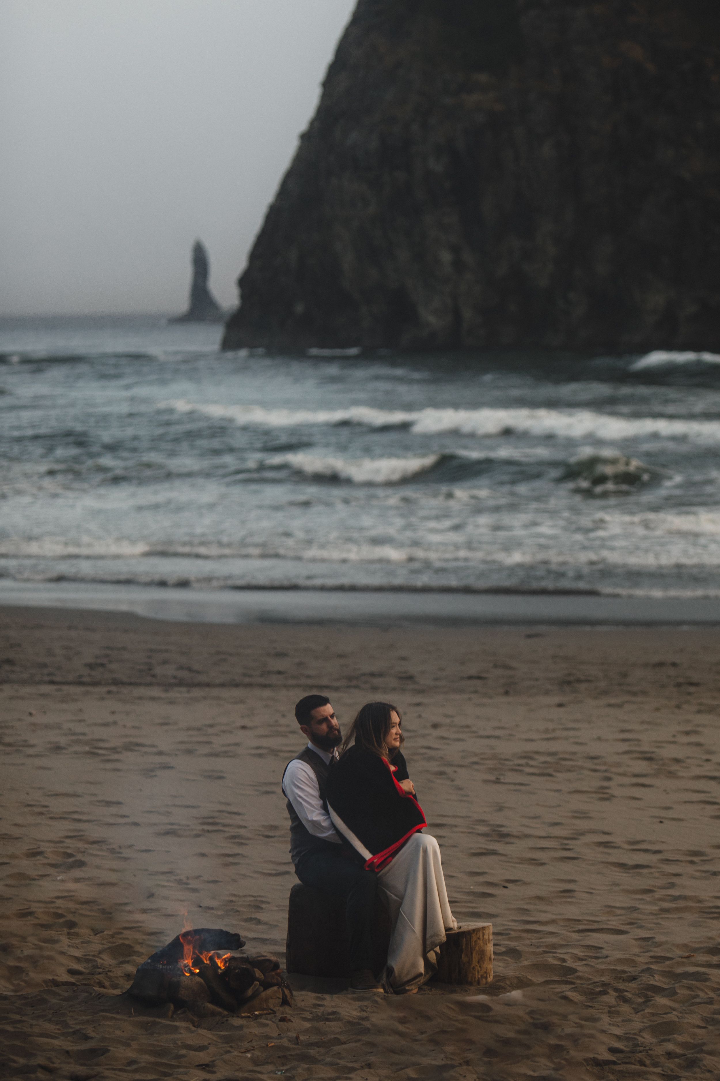 Couple sits by campfire on misty beach with dramatic cliffs and sea stack in background at sunset.