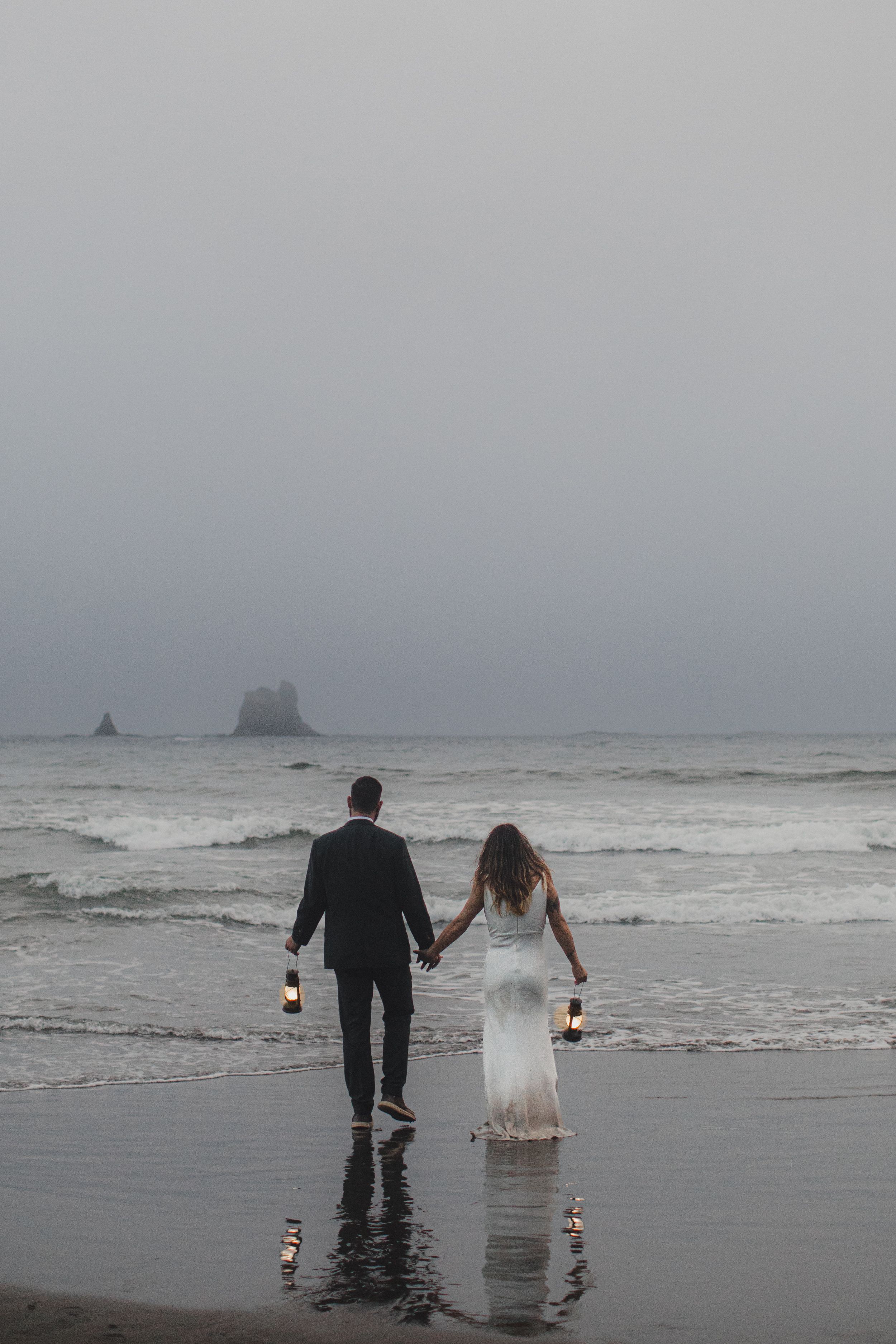 A newlywed couple holding lanterns walks along a misty beach shore with sea stacks visible in the distance.