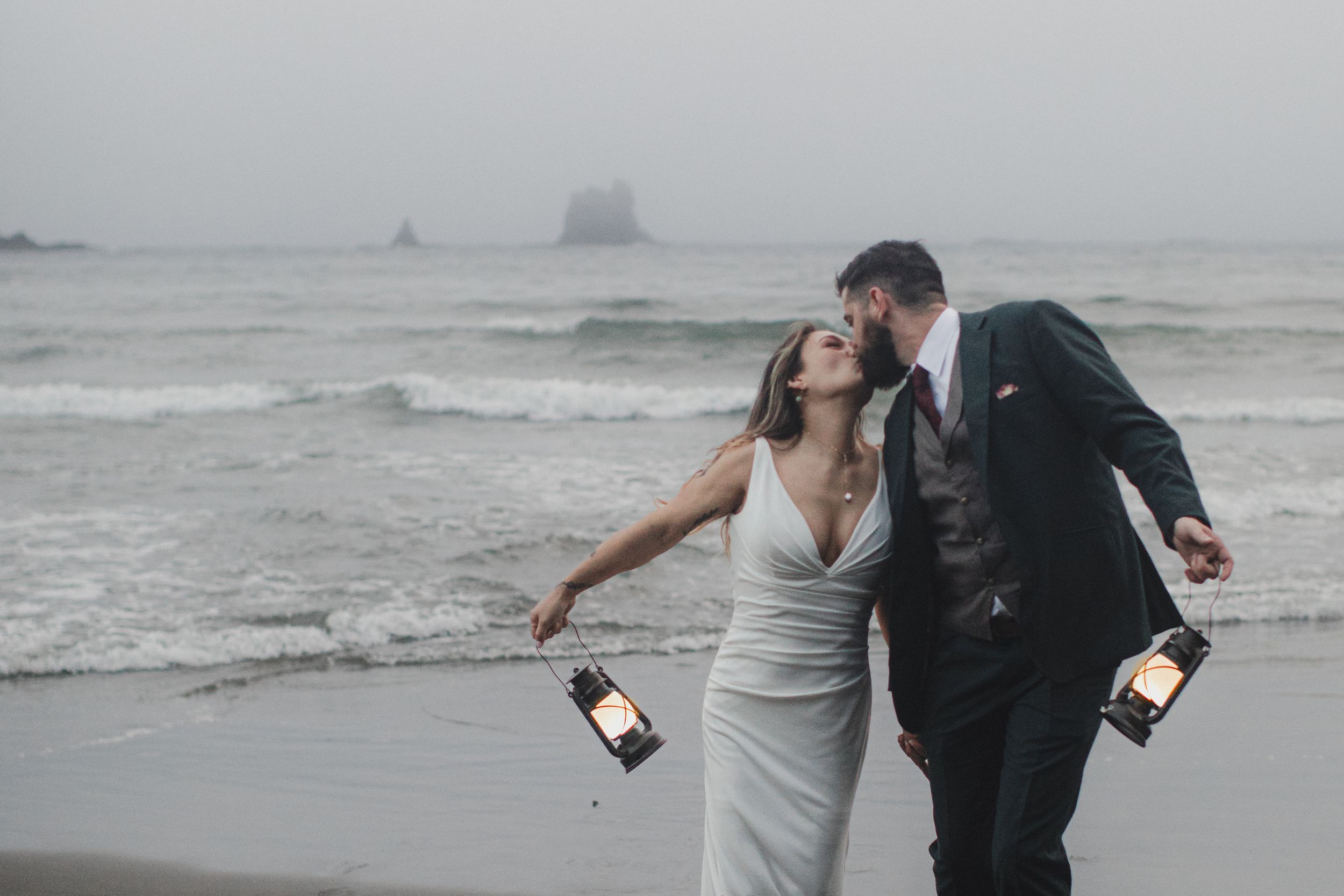 A couple embraces and shares drinks together on a foggy beach with crashing waves in the background.