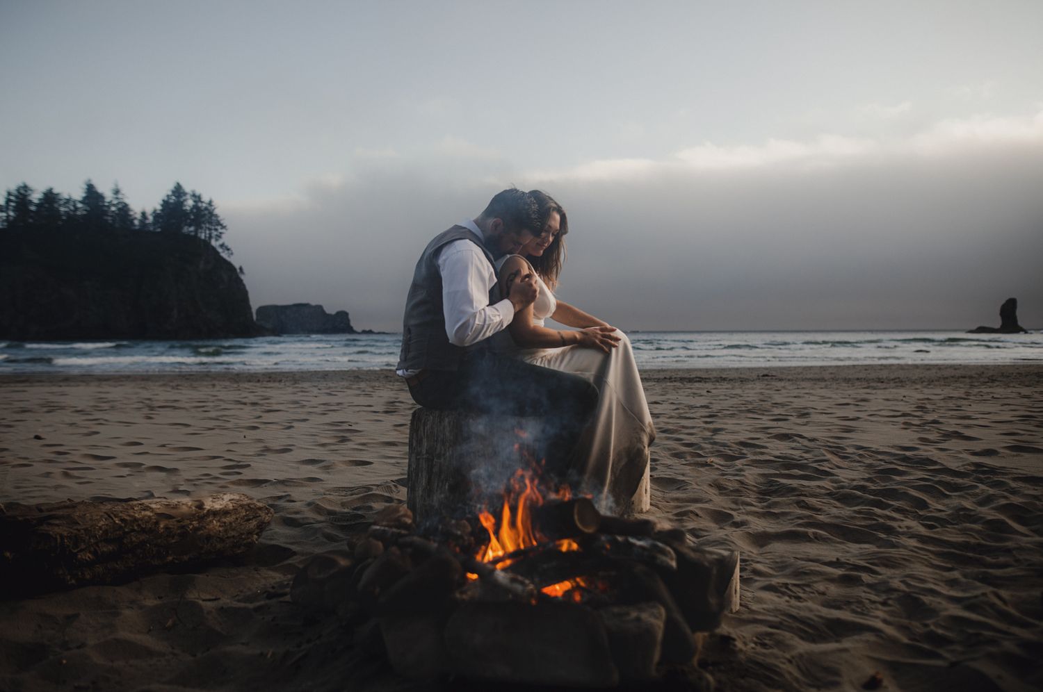 A campfire warms two people on a misty beach at dusk with evergreen trees and sea stacks in the background.