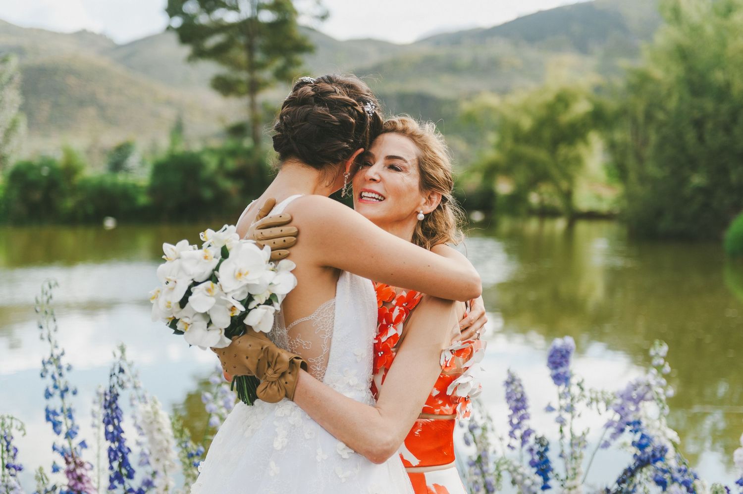 Two people embrace warmly by a lake with mountains and purple lupines in the foreground.