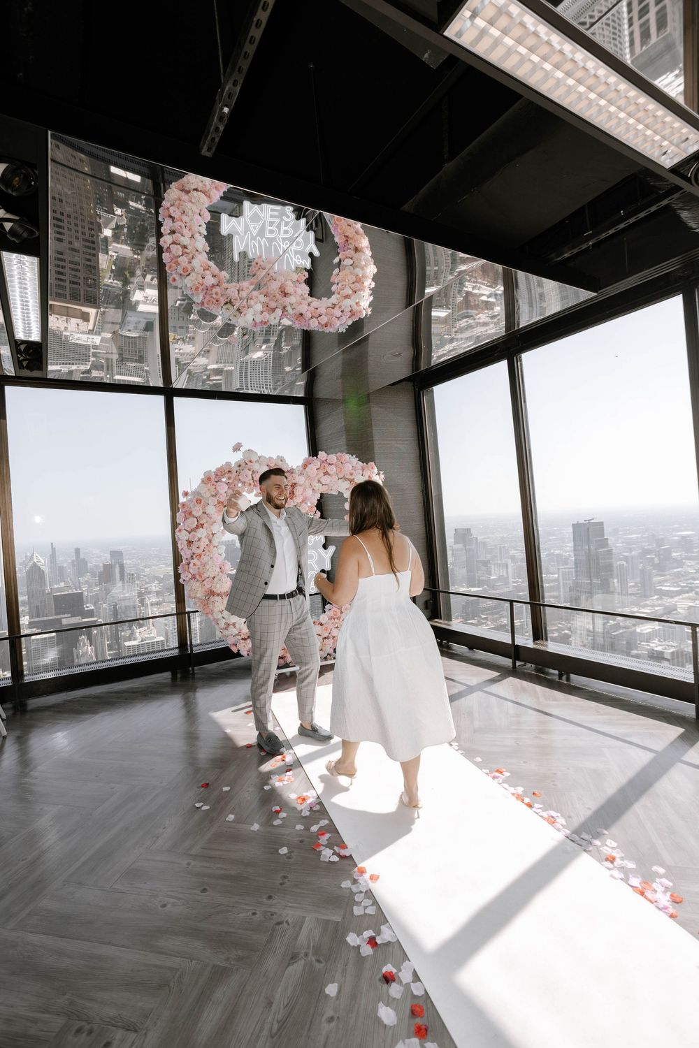 Five sequential photos capturing a romantic proposal in a modern high-rise space with pink floral decorations.