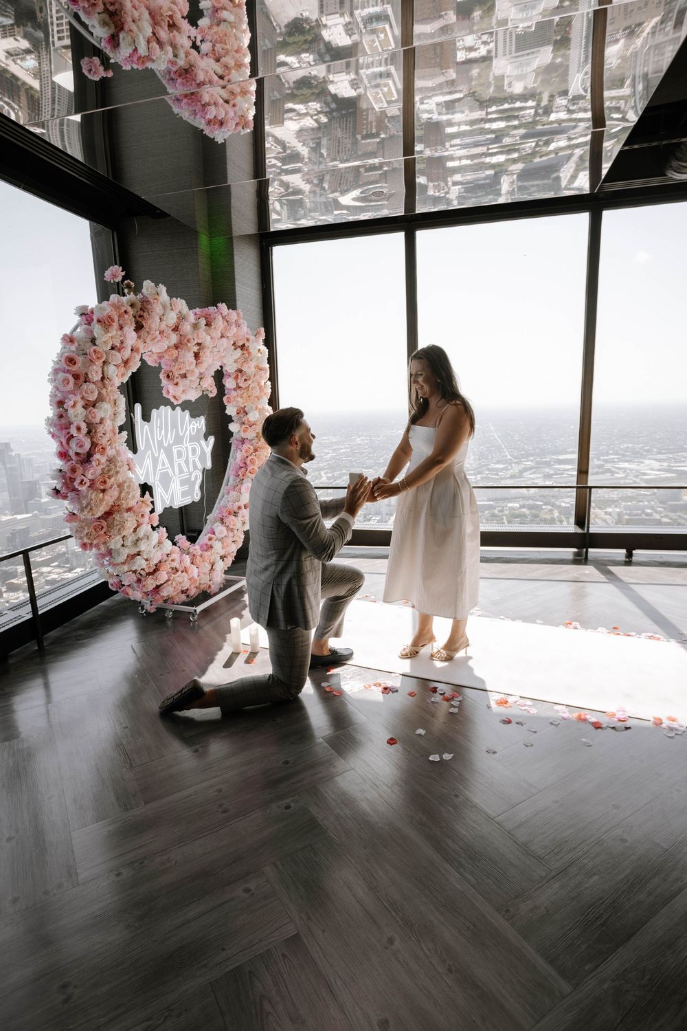 A romantic engagement moment in front of a pink floral heart arch with panoramic city views in the background.