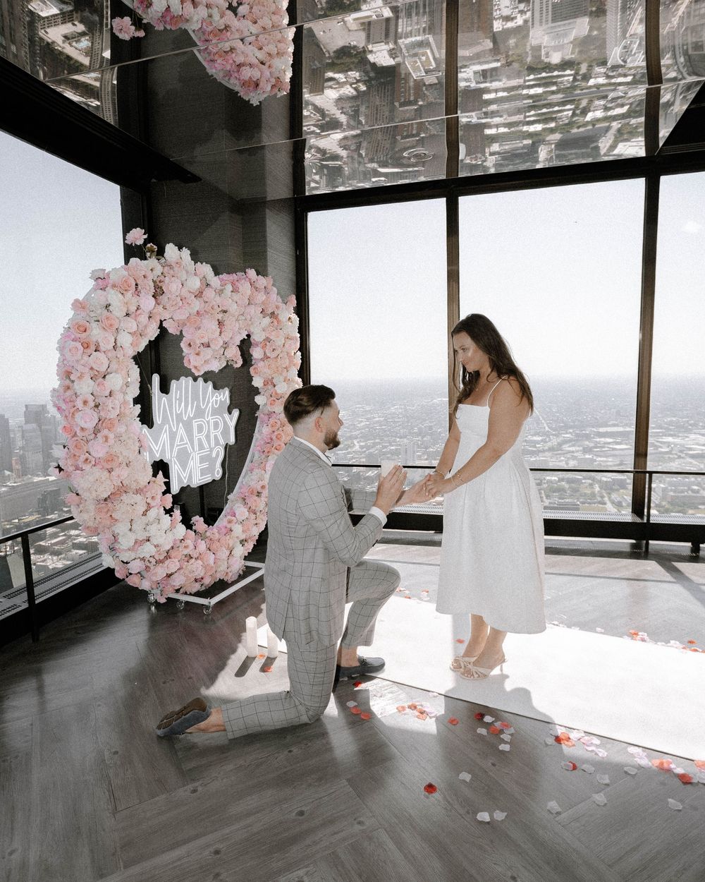 A romantic proposal scene with pink cherry blossom decor and city views through large windows.