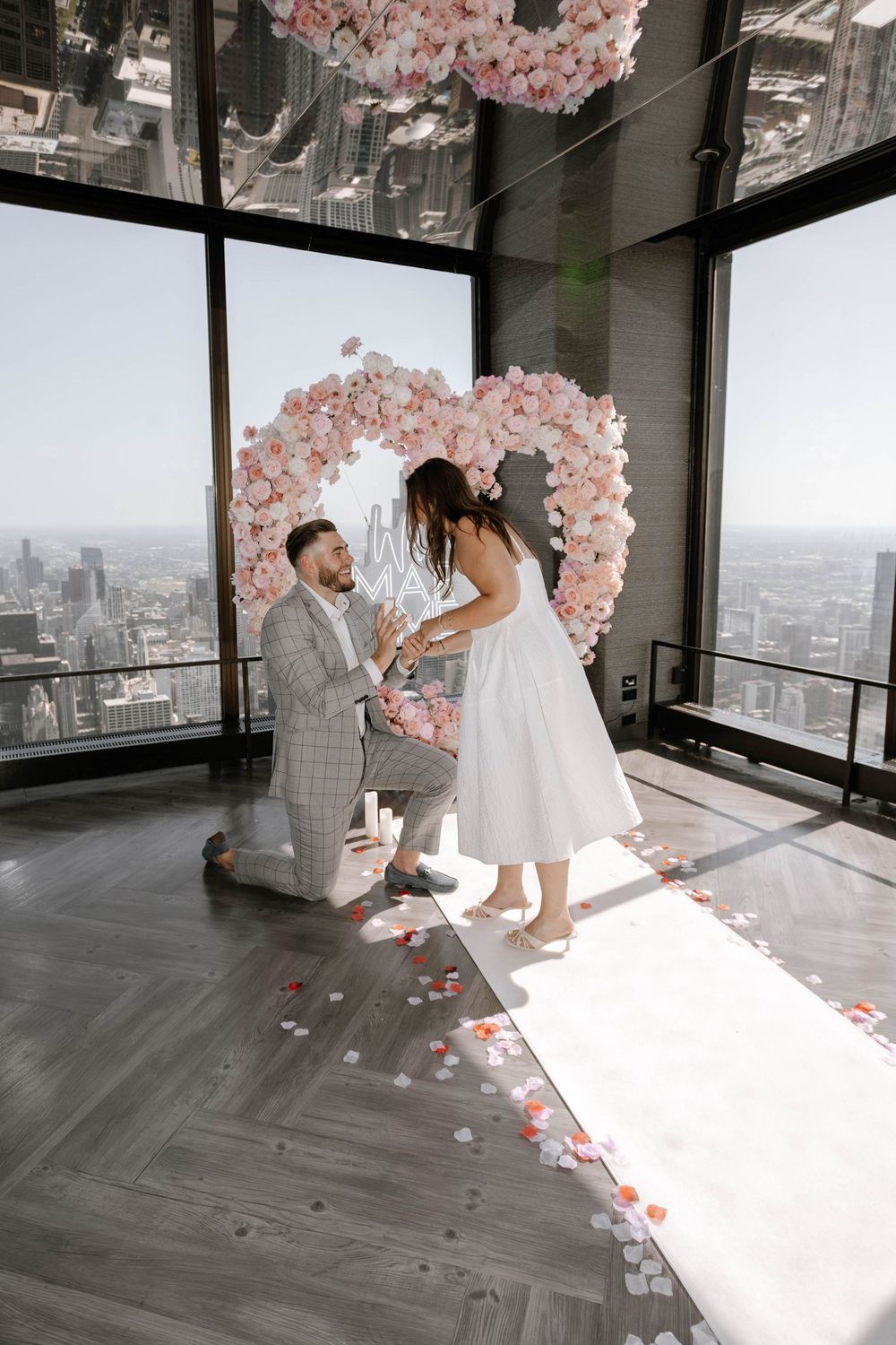 Marriage proposal scene with pink floral heart arch against cityscape backdrop in modern high-rise setting.