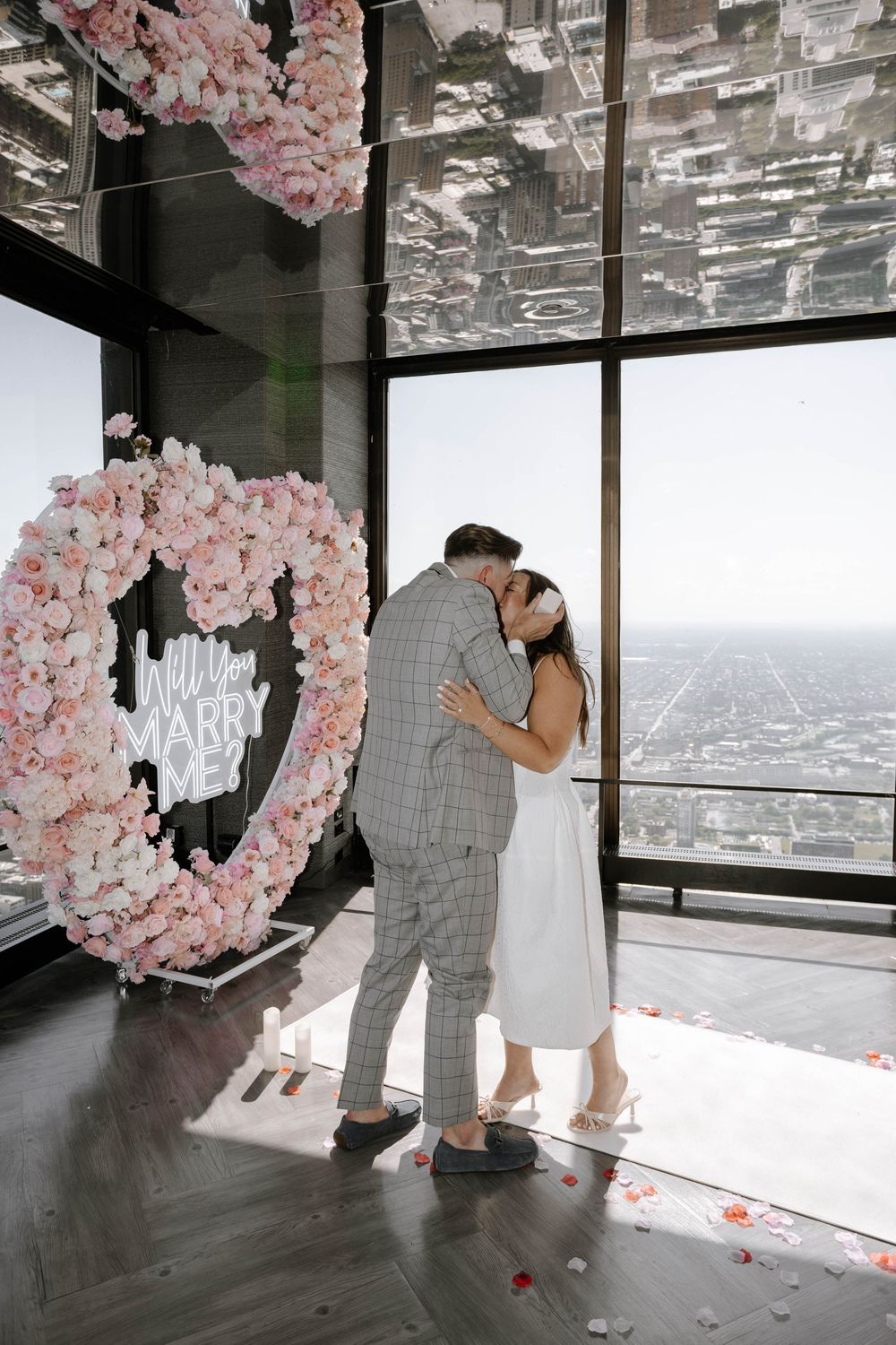Romantic moment under pink floral heart arch with cityscape views through floor-to-ceiling windows.