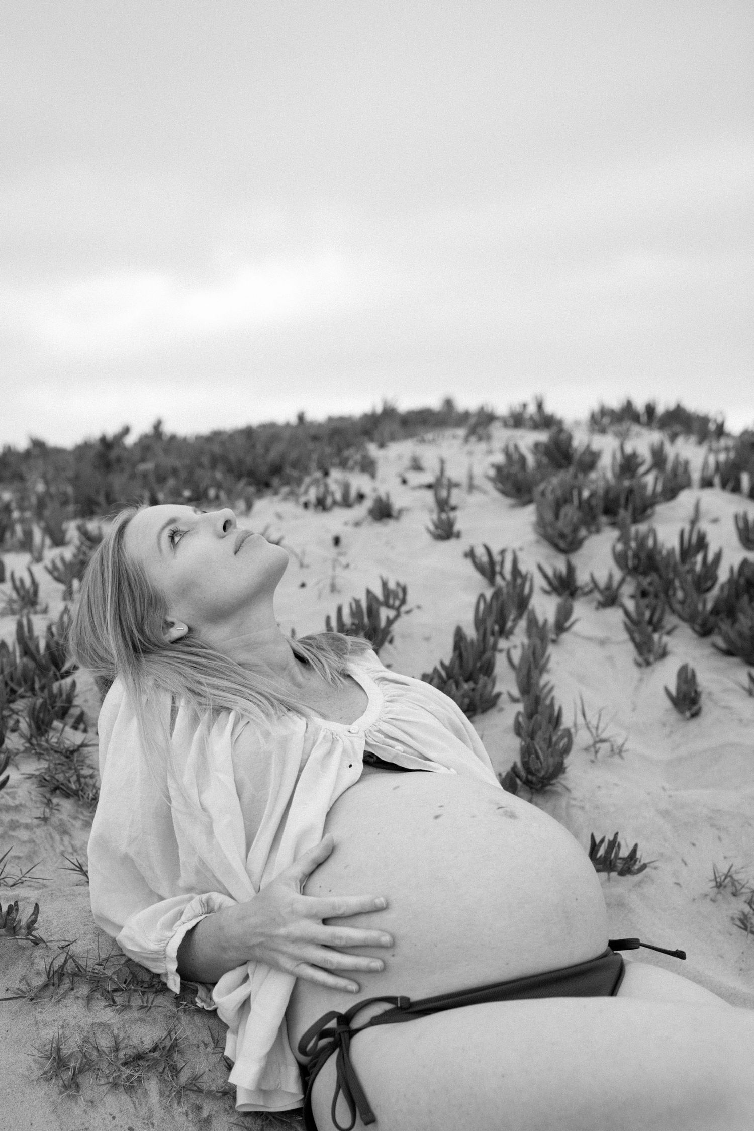 Black and white artistic photograph shows a person lying peacefully in desert sand among small desert plants.