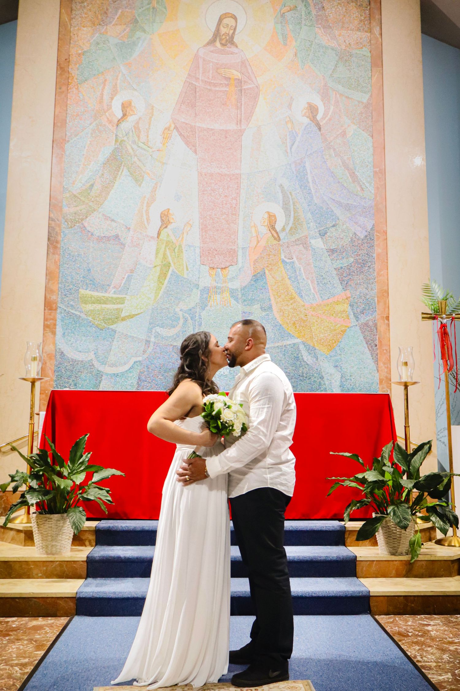 Bride and groom share romantic moment in front of colorful church mural and red altar decorations.