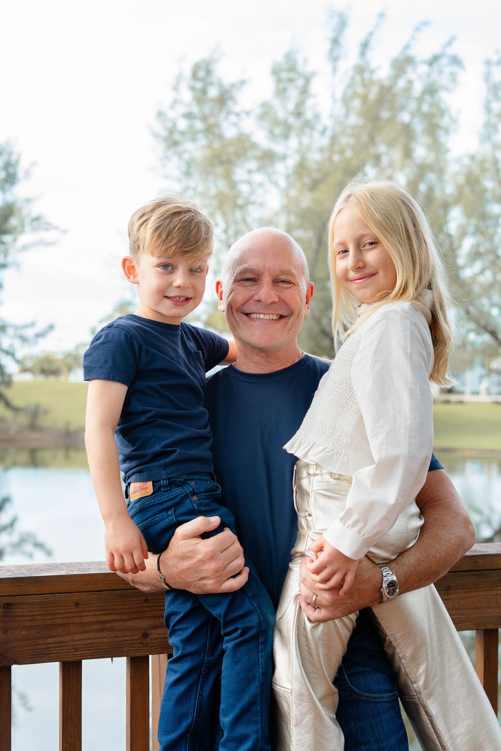 Candid outdoor family photoshoot showing three people laughing and embracing on a wooden deck by the water.